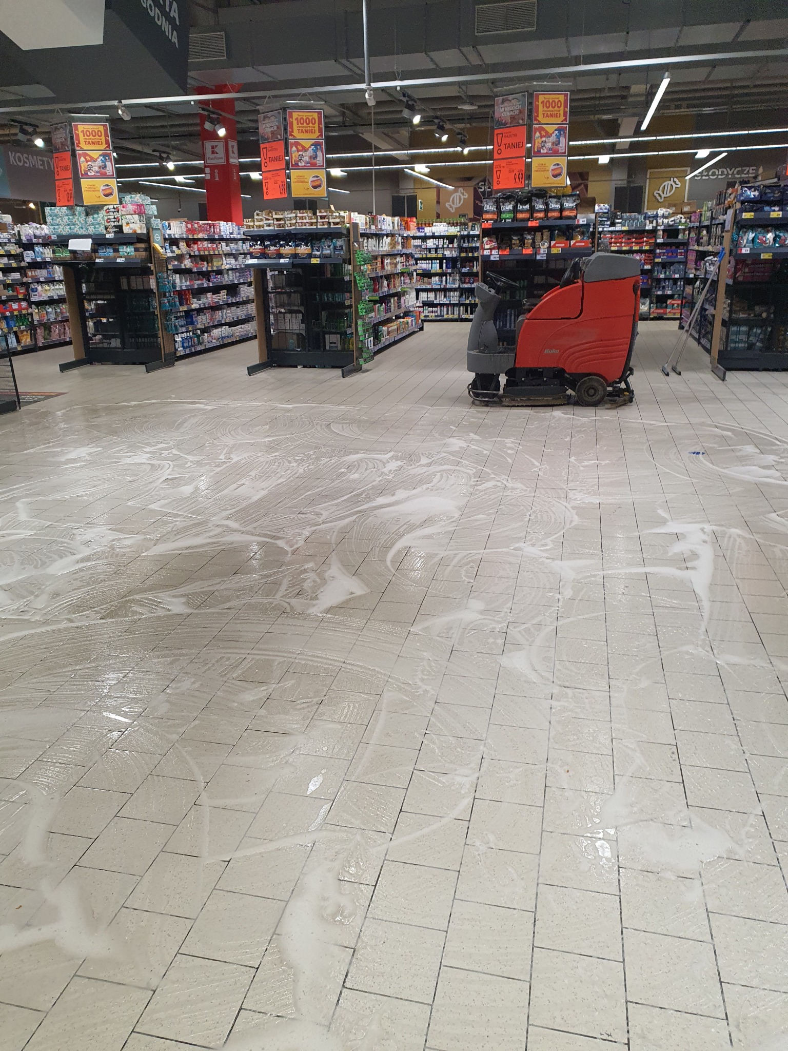 Czerwona maszyna czyszcząca marki Hako pozostawia ślady piany na jasnej, kwadratowej podłodze w supermarkecie między regałami z towarem.