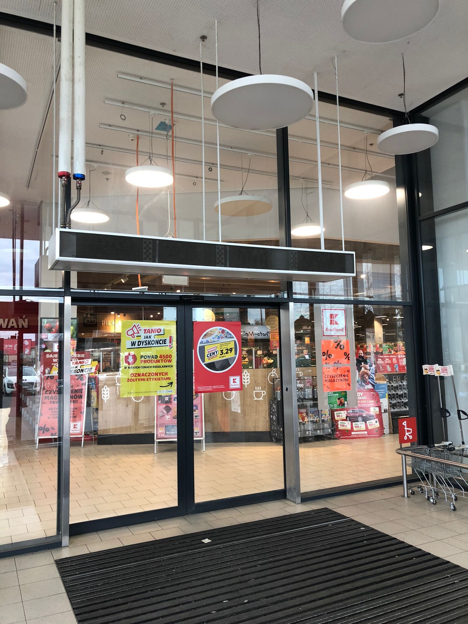 Wejście do sklepu Kaufland z widocznymi plakatami promocyjnymi na szklanych drzwiach i matą wejściową na podłodze.