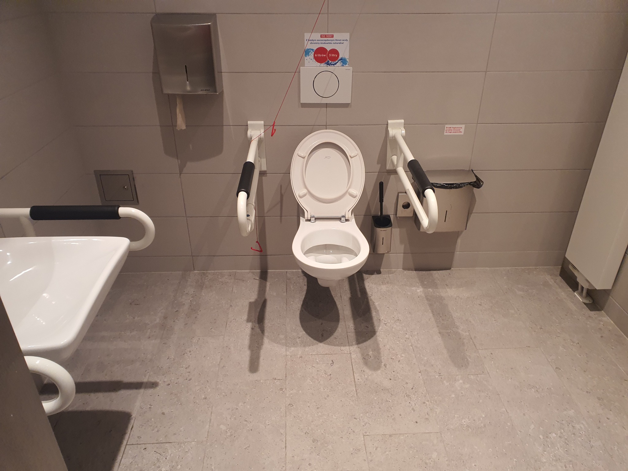 Toaleta dla niepełnosprawnych z uchwytami, dozownikiem papieru, koszem na śmieci i szczotką do WC na tle szarych płytek.