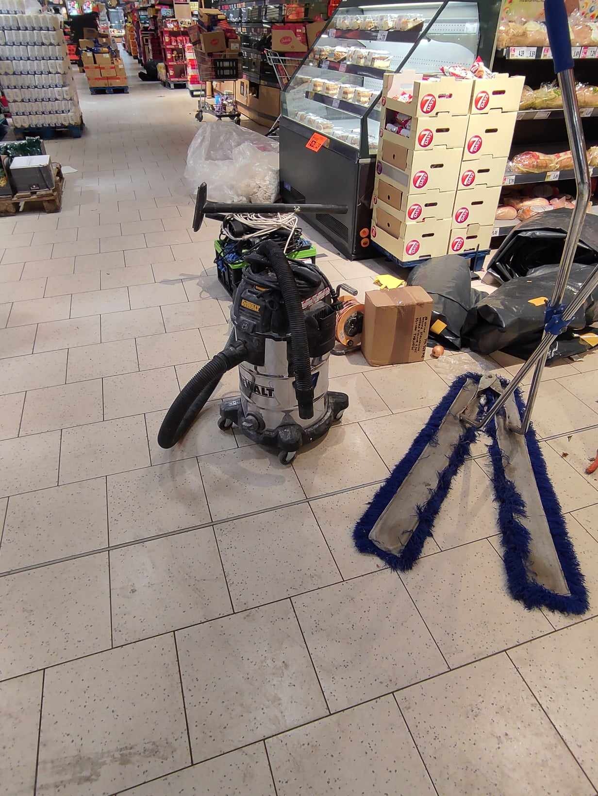 Profesjonalny odkurzacz przemysłowy DeWalt na kółkach, mop z niebieskimi frędzlami i inne akcesoria na kafelkowej podłodze w supermarkecie, sugerujące prace porządkowe.