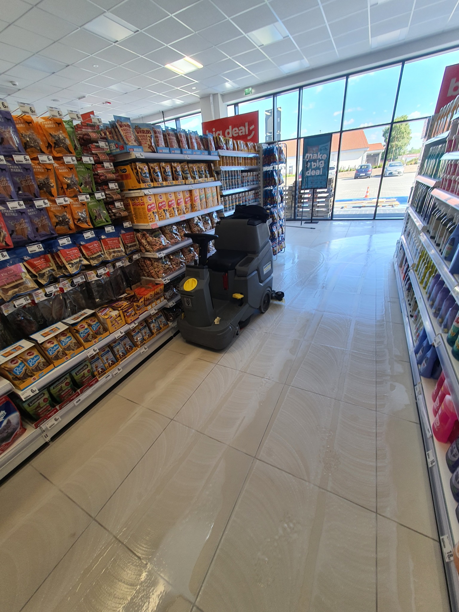 Automat do mycia podłóg w supermarkecie, czyszczący kafelki pomiędzy półkami z towarem, widoczne ślady wilgoci po myciu.