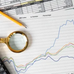 Wykres liniowy z kolorowymi liniami trendów, kalkulator, ołówek i lupa leżą na dokumencie z danymi finansowymi.