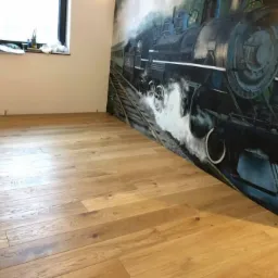 Jasne, drewniane deski podłogowe ułożone w pomieszczeniu z oknem i muralem przedstawiającym parowóz.