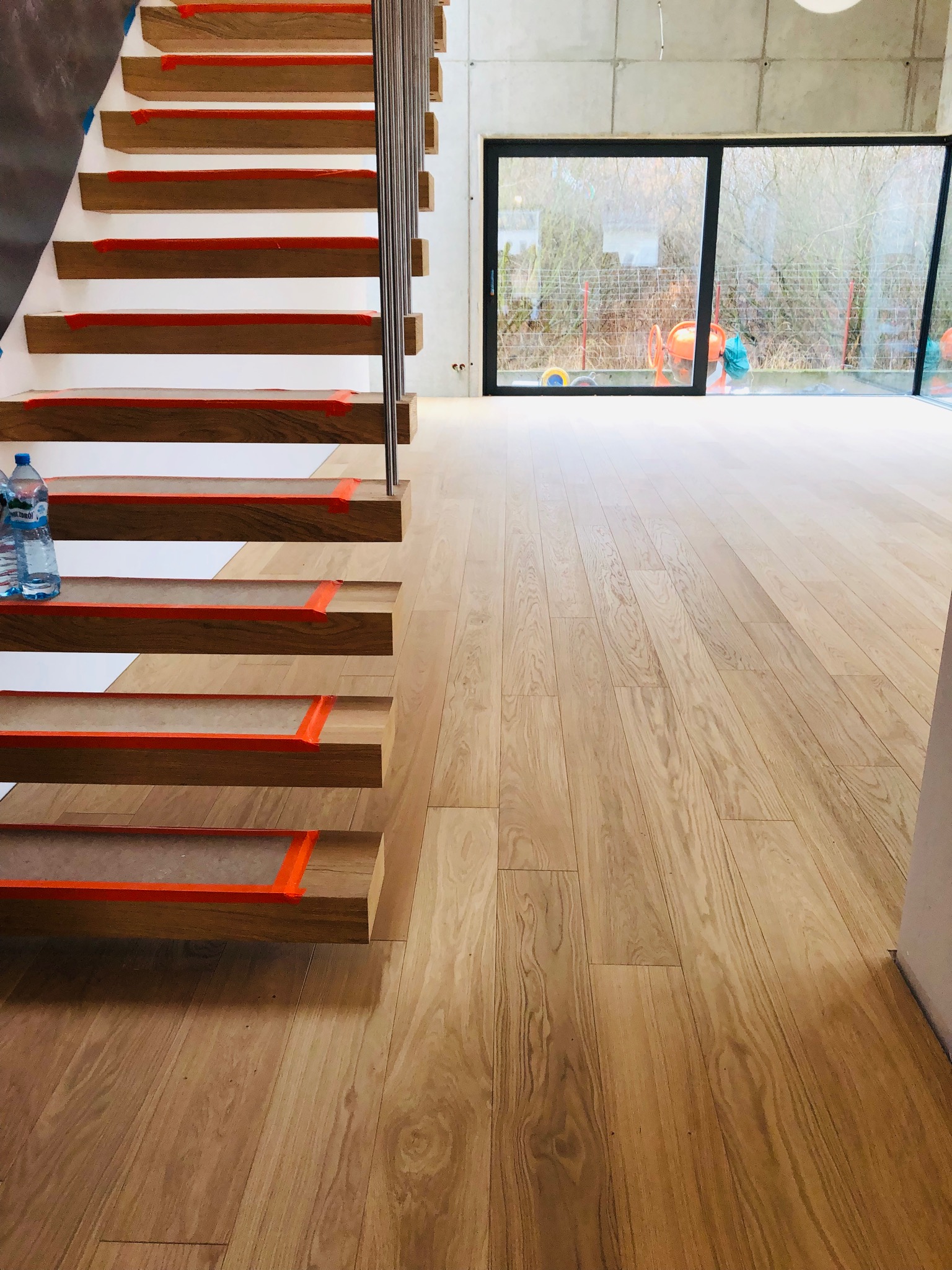Jasny parkiet z drewna dębowego w nowoczesnym wnętrzu, widoczne schody z drewnianymi stopniami zabezpieczonymi pomarańczową taśmą, butelki wody na pierwszym stopniu, duże okno przesuwne z widokiem...