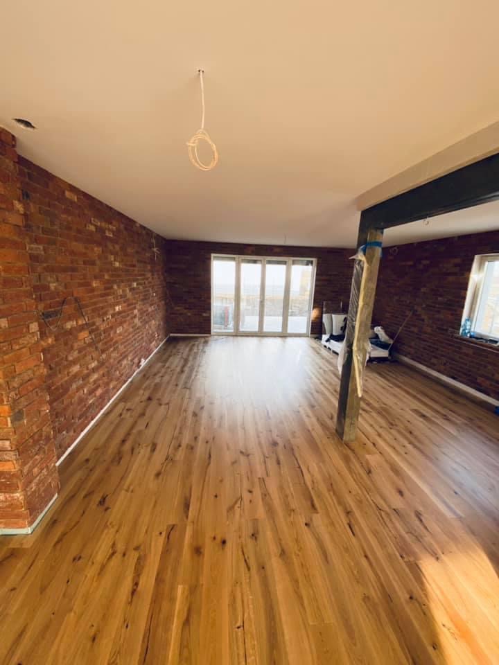 Wnętrze pomieszczenia z nowo ułożoną, lakierowaną podłogą z naturalnego drewna, ceglana ściana z widocznymi śladami prac remontowych, niezabezpieczona instalacja elektryczna zwisająca z sufitu.