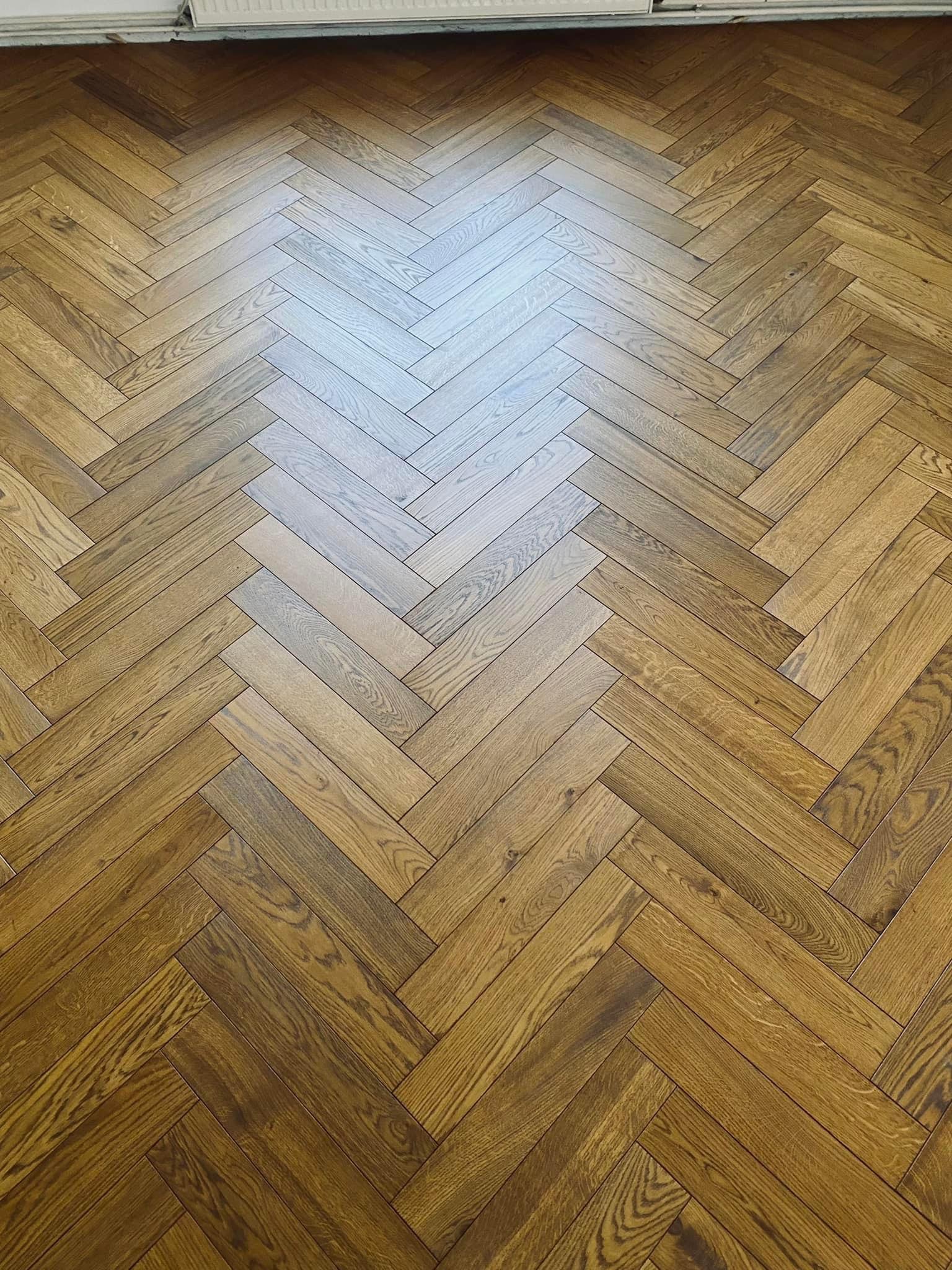 Jasny parkiet ułożony we wzór jodełki francuskiej, z widocznymi słojami drewna i subtelnym połyskiem, oświetlony naturalnym światłem.