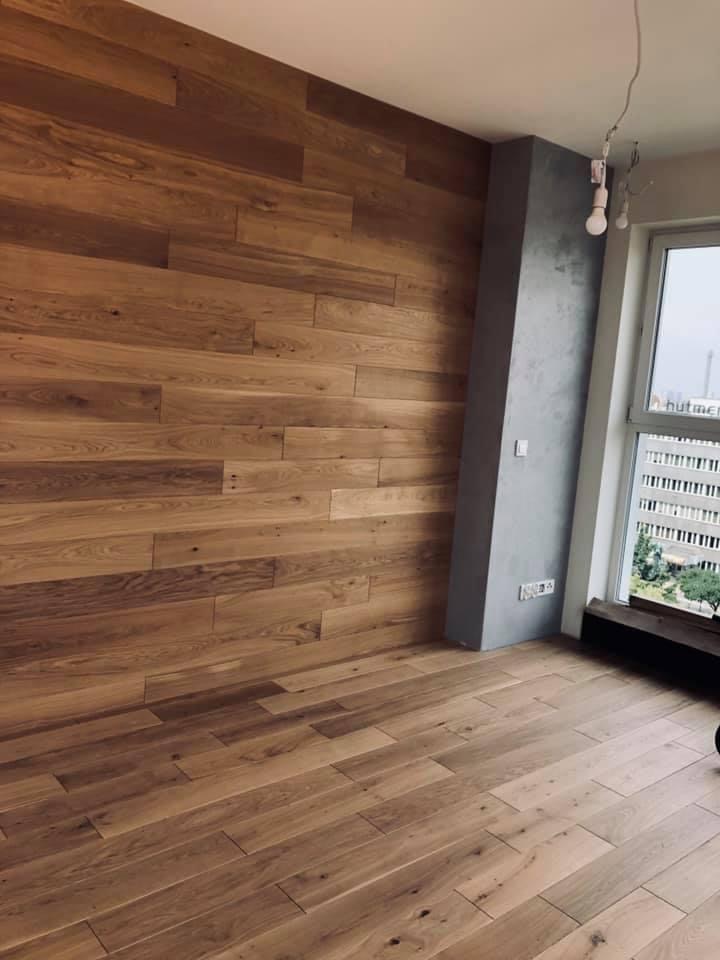 Wnętrze z drewnianą ścianą i podłogą z jasnego drewna, widok na szary filar i okno z widokiem na budynek biurowy.