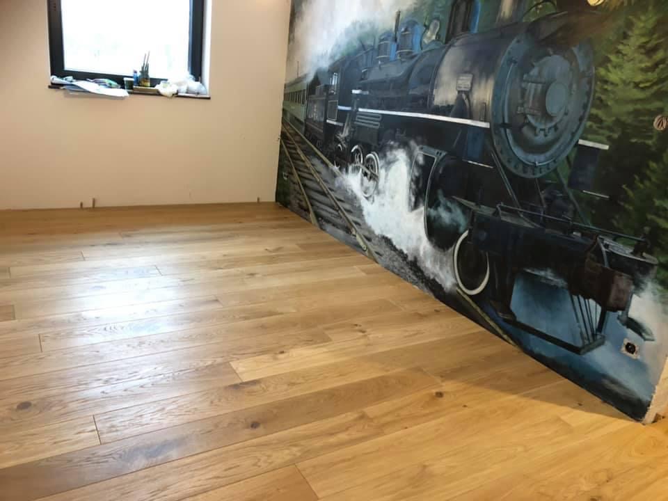 Jasne, drewniane deski podłogowe ułożone w pomieszczeniu z oknem i muralem przedstawiającym parowóz.