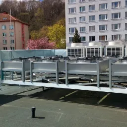 Rząd jednostek zewnętrznych klimatyzacji przemysłowej na dachu budynku, widok z perspektywy dachu, w tle budynki mieszkalne i drzewa.