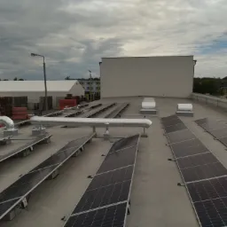 Widok z góry na panele fotowoltaiczne i elementy wentylacji na płaskim dachu budynku w Straszynie, pochmurne niebo w tle.