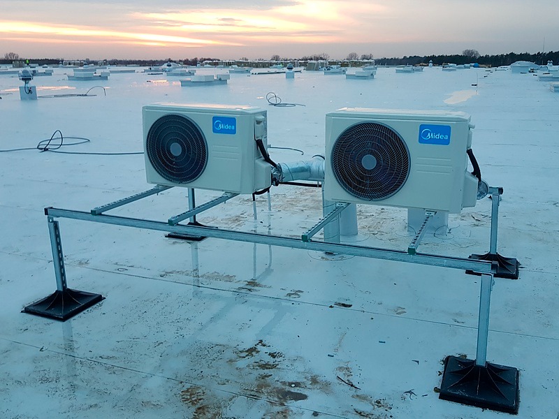 Dwa klimatyzatory Midea zamontowane na metalowej konstrukcji na płaskim, białym dachu budynku komercyjnego, widoczne inne elementy instalacji na dachu i zachodzące słońce w tle.