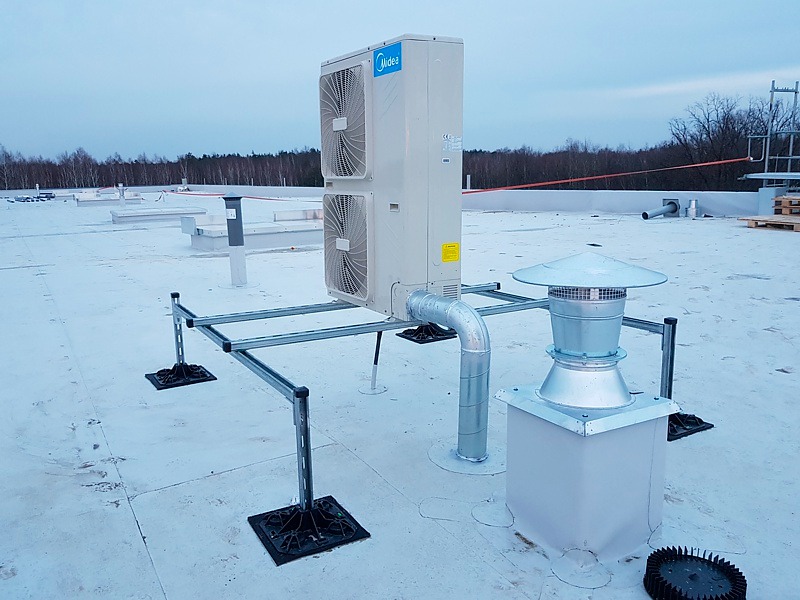 Klimatyzator Midea na płaskim dachu budynku komercyjnego, zamontowany na metalowej konstrukcji wsporczej, obok komina wentylacyjnego z daszkiem.