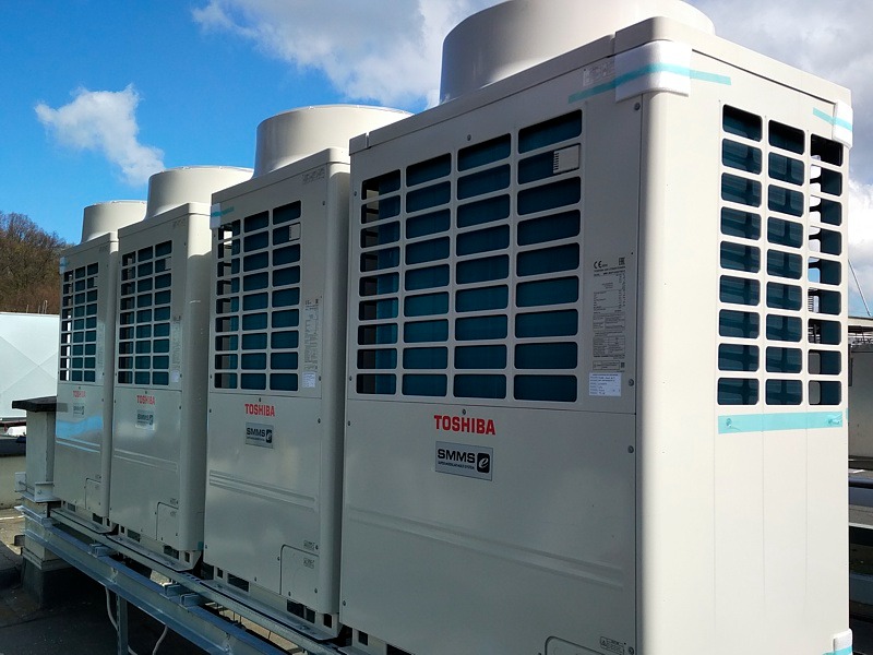 Rząd czterech identycznych, białych jednostek zewnętrznych klimatyzacji Toshiba SMMS ustawionych na metalowej konstrukcji na dachu budynku, widoczne niebieskie niebo z chmurami w tle.