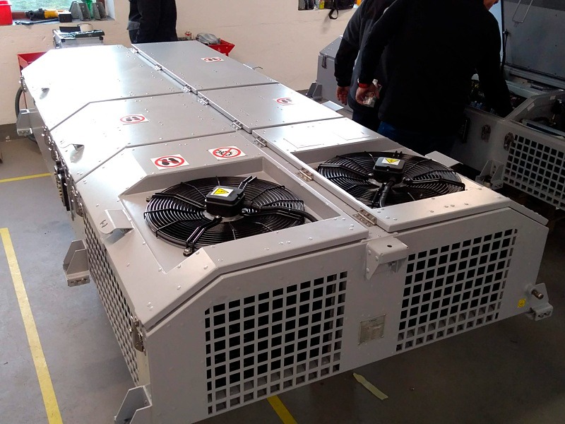 Duża, przemysłowa jednostka klimatyzacyjna z dwoma wentylatorami i znakami ostrzegawczymi, w trakcie montażu lub serwisu, widoczne osoby obsługujące urządzenie.