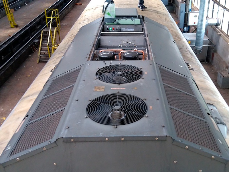 Widok z góry na klimatyzację przemysłową zamontowaną na dachu pojazdu szynowego, widoczne wentylatory, kratki wentylacyjne i elementy instalacji chłodniczej.