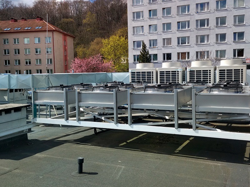 Rząd jednostek zewnętrznych klimatyzacji przemysłowej na dachu budynku, widok z perspektywy dachu, w tle budynki mieszkalne i drzewa.