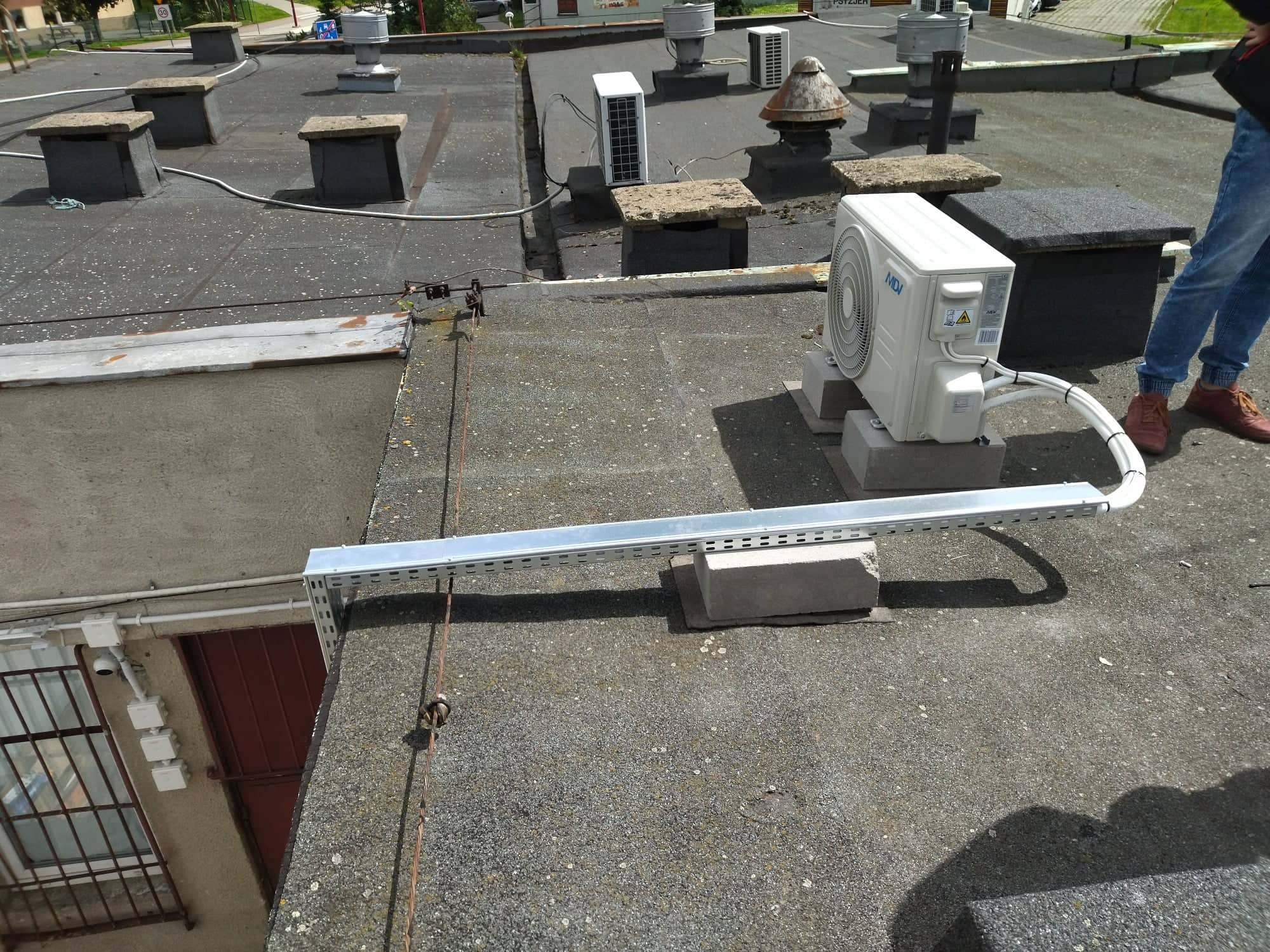 Jednostka zewnętrzna klimatyzacji umieszczona na betonowych bloczkach na płaskim, bitumicznym dachu budynku komercyjnego, widoczne kominy wentylacyjne i fragment instalacji elektrycznej.