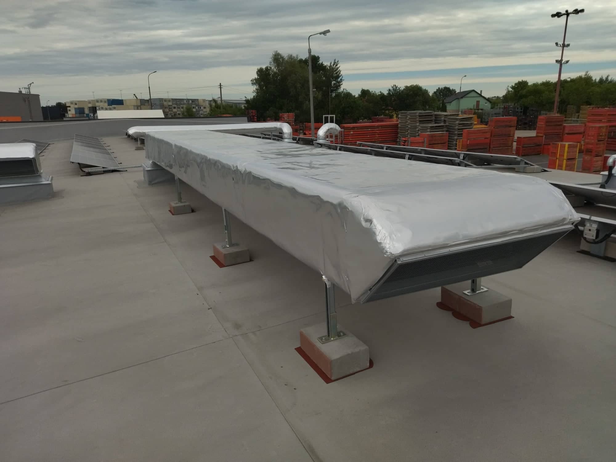 Długi, prostokątny kanał wentylacyjny w srebrnej izolacji na dachu budynku komercyjnego, wsparty na betonowych podstawach z czerwonymi podkładkami antywibracyjnymi.