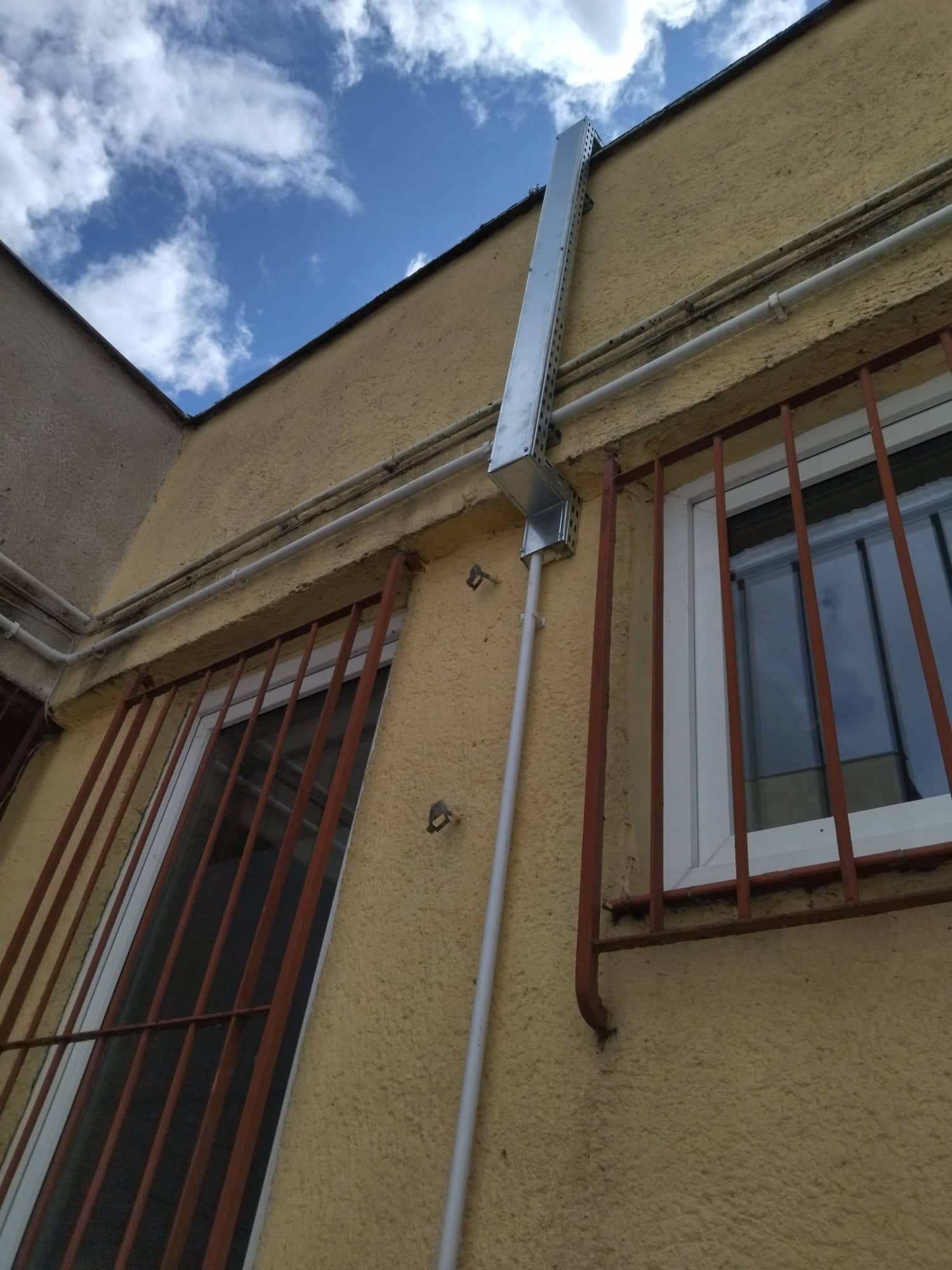 Zewnętrzna instalacja wentylacyjna w metalowej osłonie, pionowy kanał wentylacyjny na elewacji budynku z oknami zabezpieczonymi kratami.