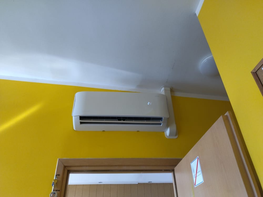 Biała klimatyzacja ścienna na żółtej ścianie, widoczny fragment drzwi z naklejką zakazu palenia, rury instalacyjne prowadzone pod sufitem.