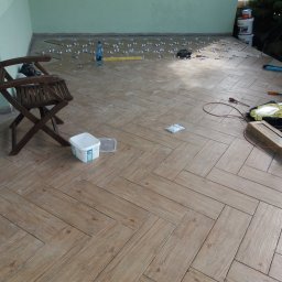 Jarek Śmigiel - Układanie płytek podłogowych imitujących drewno na balkonie w jodełkę, widoczne narzędzia, poziomica i zapas płytek z krzyżykami dystansowymi.