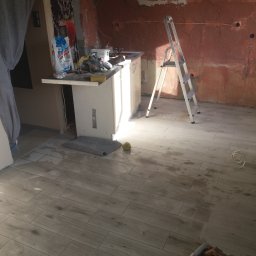 Jarek Śmigiel - Ułożone płytki podłogowe imitujące drewno w trakcie remontu kuchni, widoczna drabina, narzędzia i resztki materiałów budowlanych na blacie, ściana w stanie surowym.