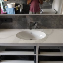 Jarek Śmigiel - Biała umywalka nablatowa z marmurowym wzorem na blacie, zamontowana na szafce z otwartymi półkami, widoczna na tle szarej ściany z marmurowym wzorem i odbiciem w lustrze, w którym widać fragment...