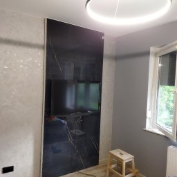 Jarek Śmigiel - Telewizor zawieszony na ścianie wyłożonej czarnymi płytkami imitującymi marmur ze złotymi żyłami, obok ściana pokryta dekoracyjnym tynkiem w odcieniach szarości, okrągła lampa sufitowa LED.