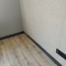 Jarek Śmigiel - Fragment wykończonego wnętrza w Gorzowie Wielkopolskim: ściana z tapetą o delikatnej strukturze, jasna podłoga laminowana, czarna listwa przypodłogowa i gniazdko elektryczne.