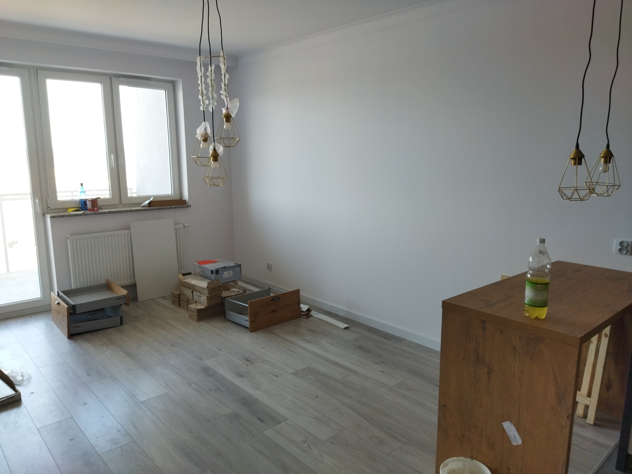 Wnętrze w trakcie wykańczania: panele podłogowe, białe ściany, wiszące lampy, części do mebli w kartonach i rozłożona wyspa kuchenna z butelką wody.
