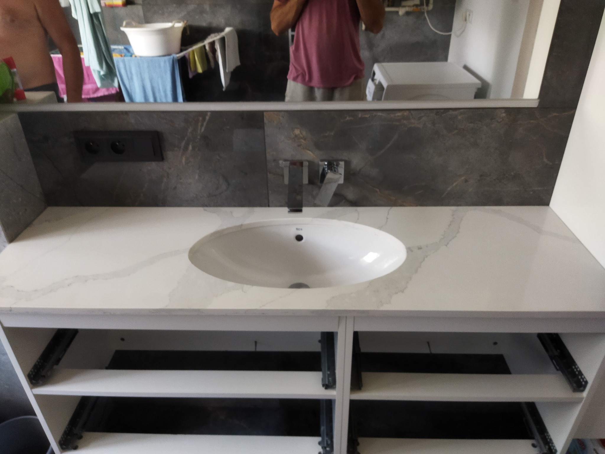 Biała umywalka nablatowa z marmurowym wzorem na blacie, zamontowana na szafce z otwartymi półkami, widoczna na tle szarej ściany z marmurowym wzorem i odbiciem w lustrze, w którym widać fragment...