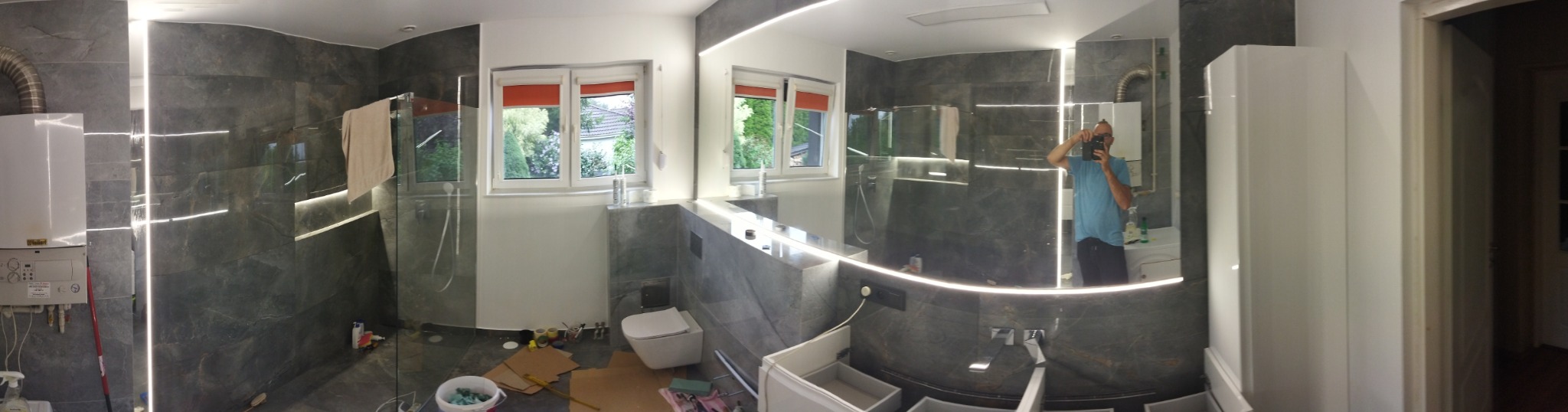 Szeroki kadr na wnętrze remontowanej łazienki z szarymi płytkami imitującymi kamień, podświetleniem LED, kabiną prysznicową typu walk-in i widocznymi elementami instalacji oraz materiałami...