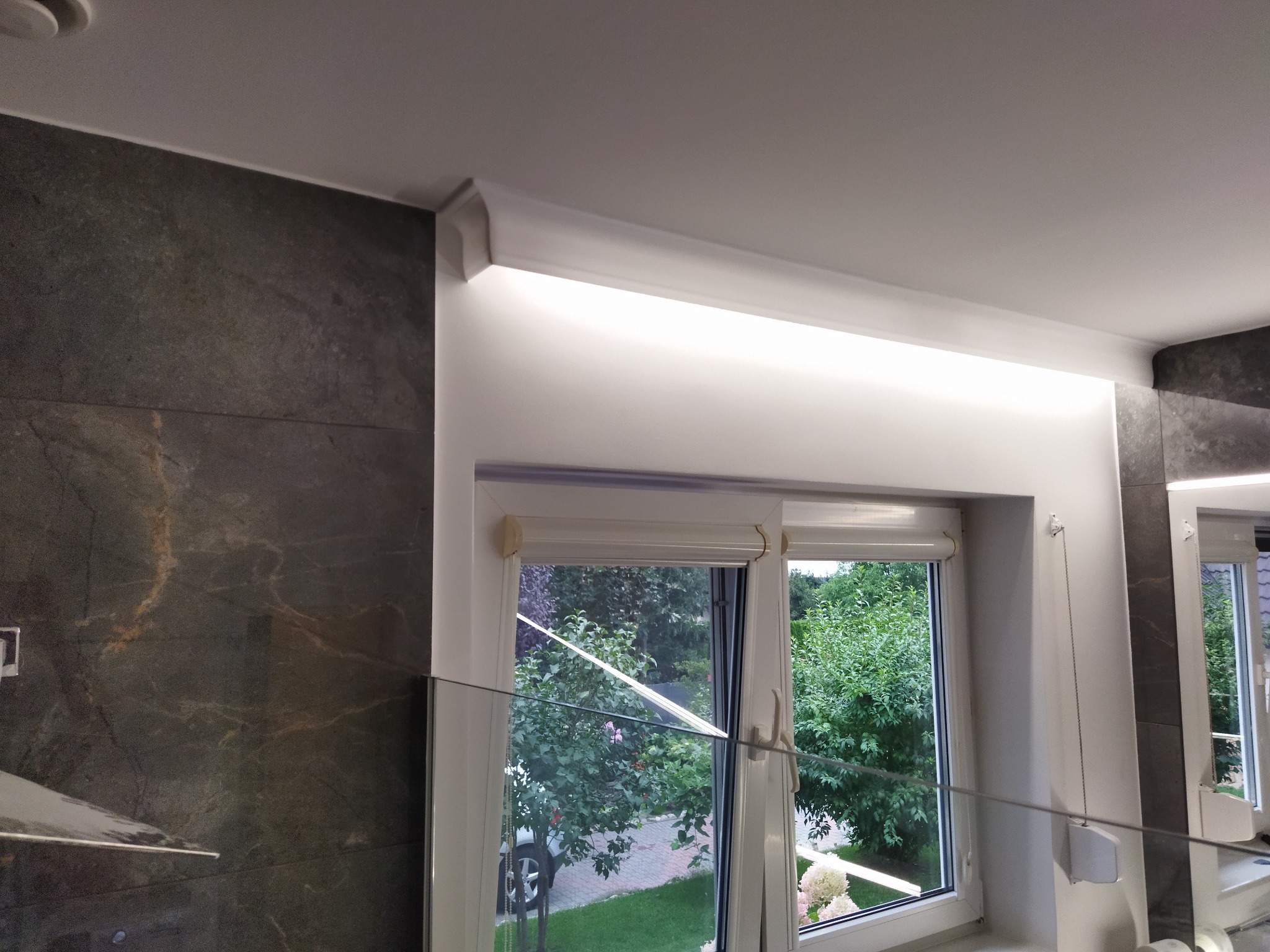 Nowoczesna łazienka z szarą płytką imitującą kamień, podświetlanym sufitem i oknem z roletami, widok z dołu.