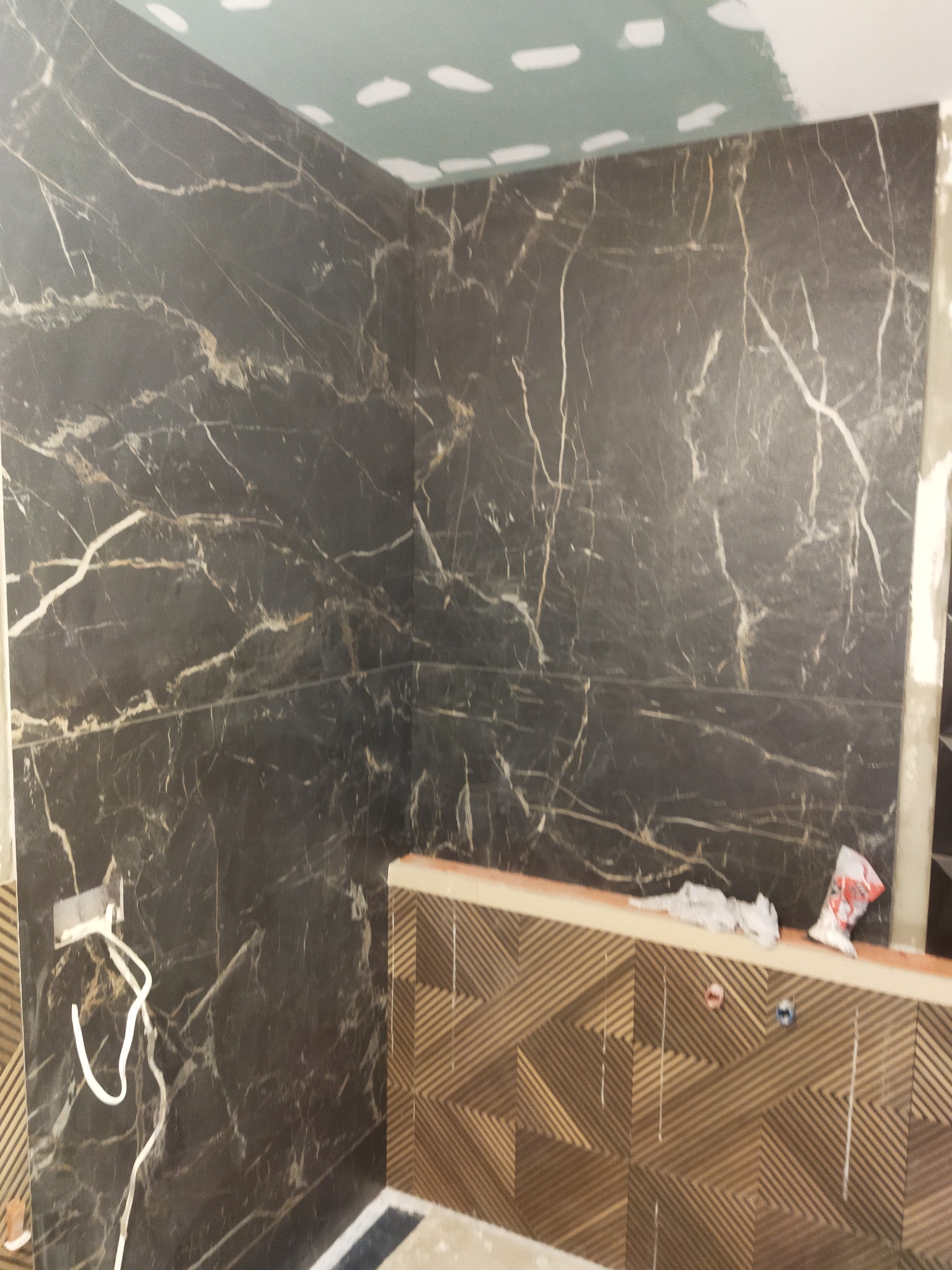 Częściowo wykończona ściana łazienki z czarnych płytek imitujących marmur z białymi żyłami, dolna część ściany wyłożona płytkami z geometrycznym wzorem, widoczne przewody elektryczne i resztki...