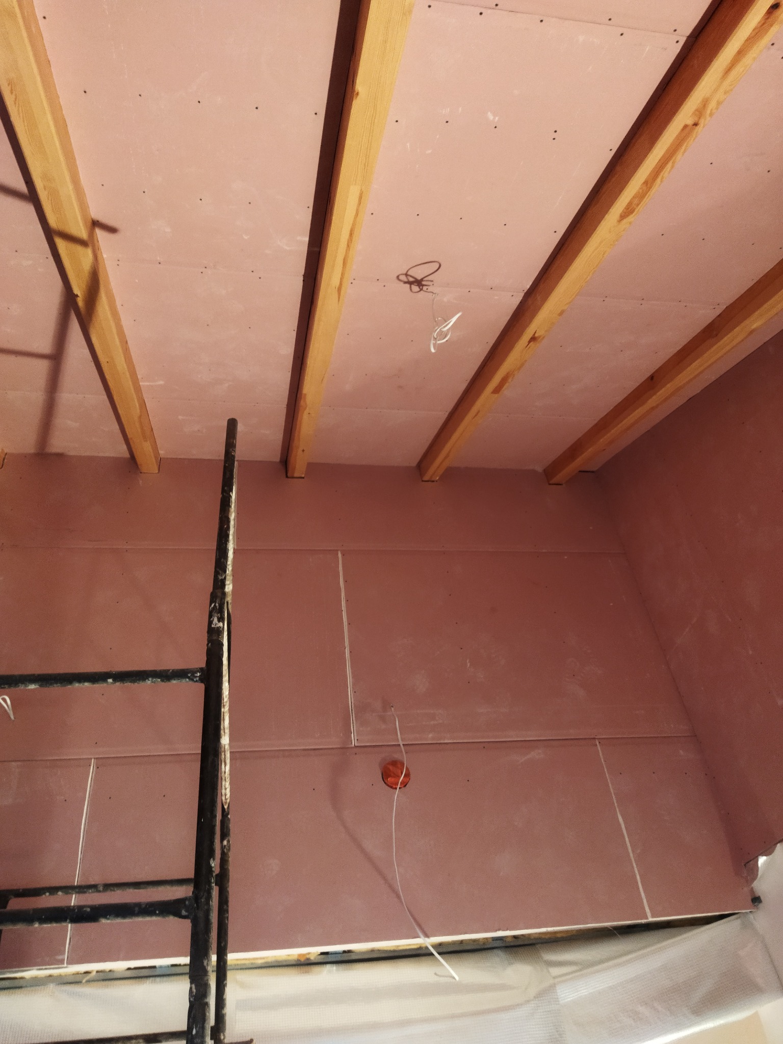 Montaż płyt kartonowo-gipsowych na ścianie i suficie, widoczne drewniane belki stropowe i fragment rusztowania.