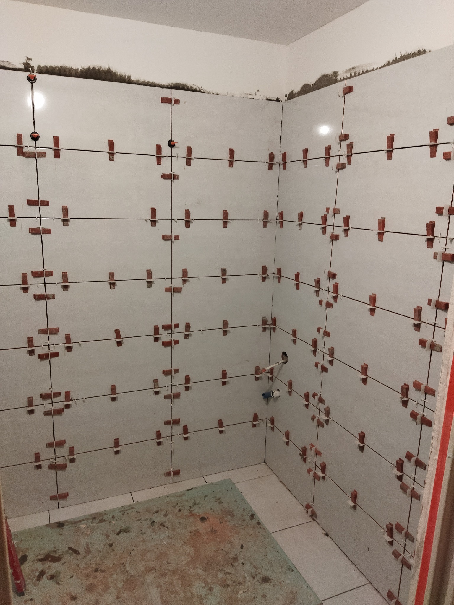 Świeżo ułożone, białe płytki w łazience, zabezpieczone krzyżykami dystansowymi i systemem poziomowania, widoczne ślady zaprawy na górze ściany.