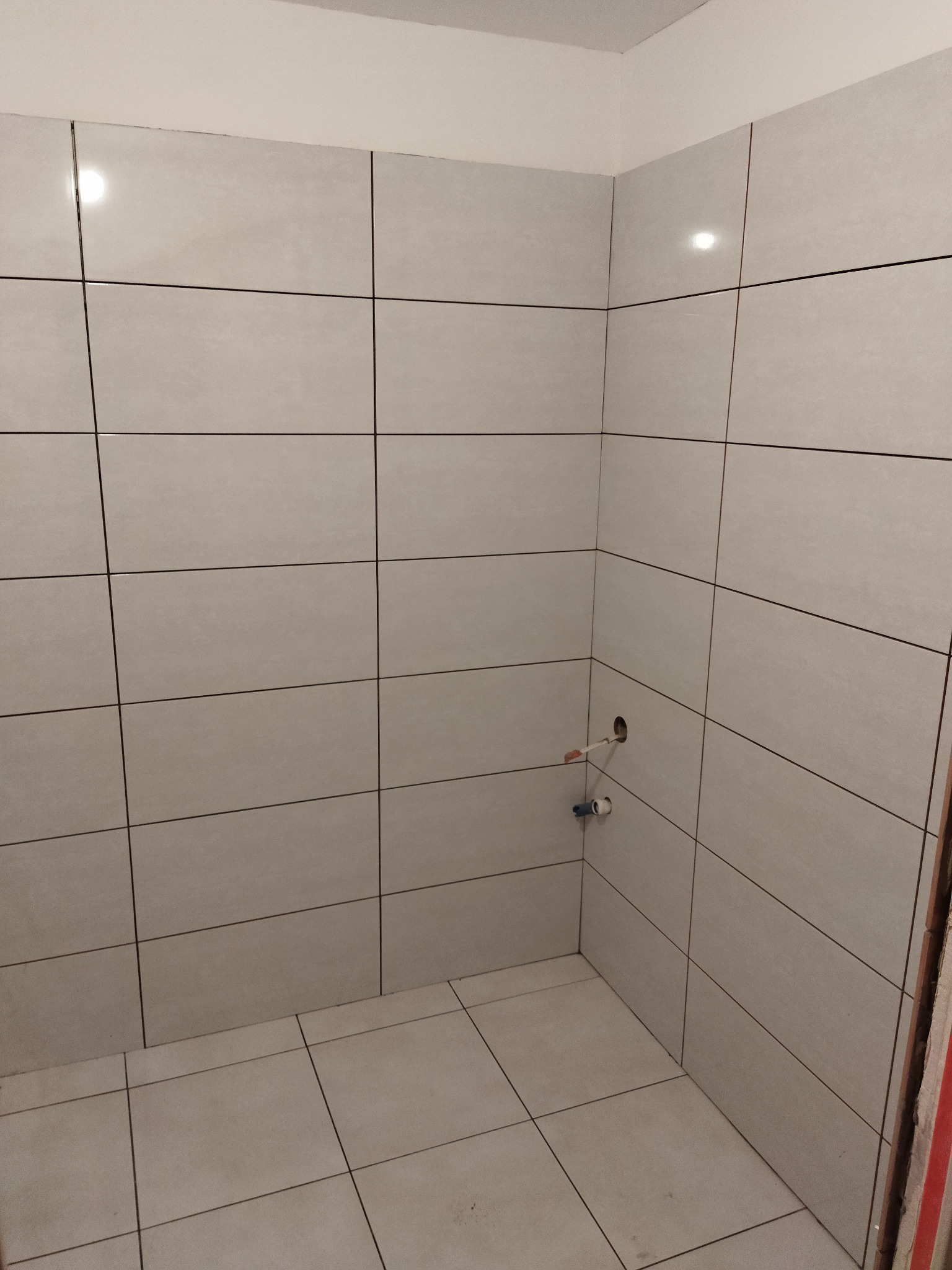 Świeżo wykafelkowana ściana i podłoga w narożnej kabinie prysznicowej, widoczne wystające rury instalacji wodnej przed montażem armatury.