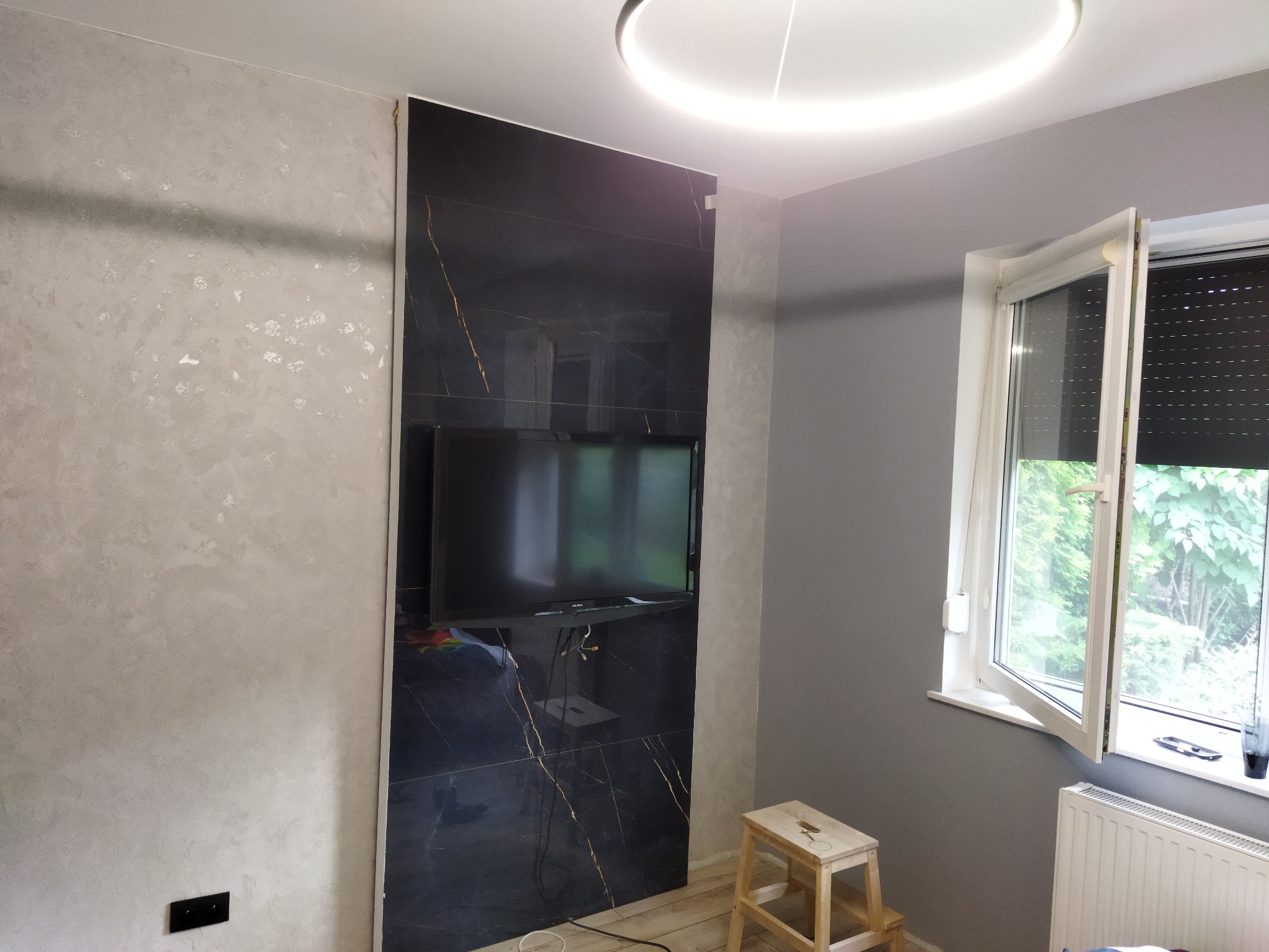 Telewizor zawieszony na ścianie wyłożonej czarnymi płytkami imitującymi marmur ze złotymi żyłami, obok ściana pokryta dekoracyjnym tynkiem w odcieniach szarości, okrągła lampa sufitowa LED.