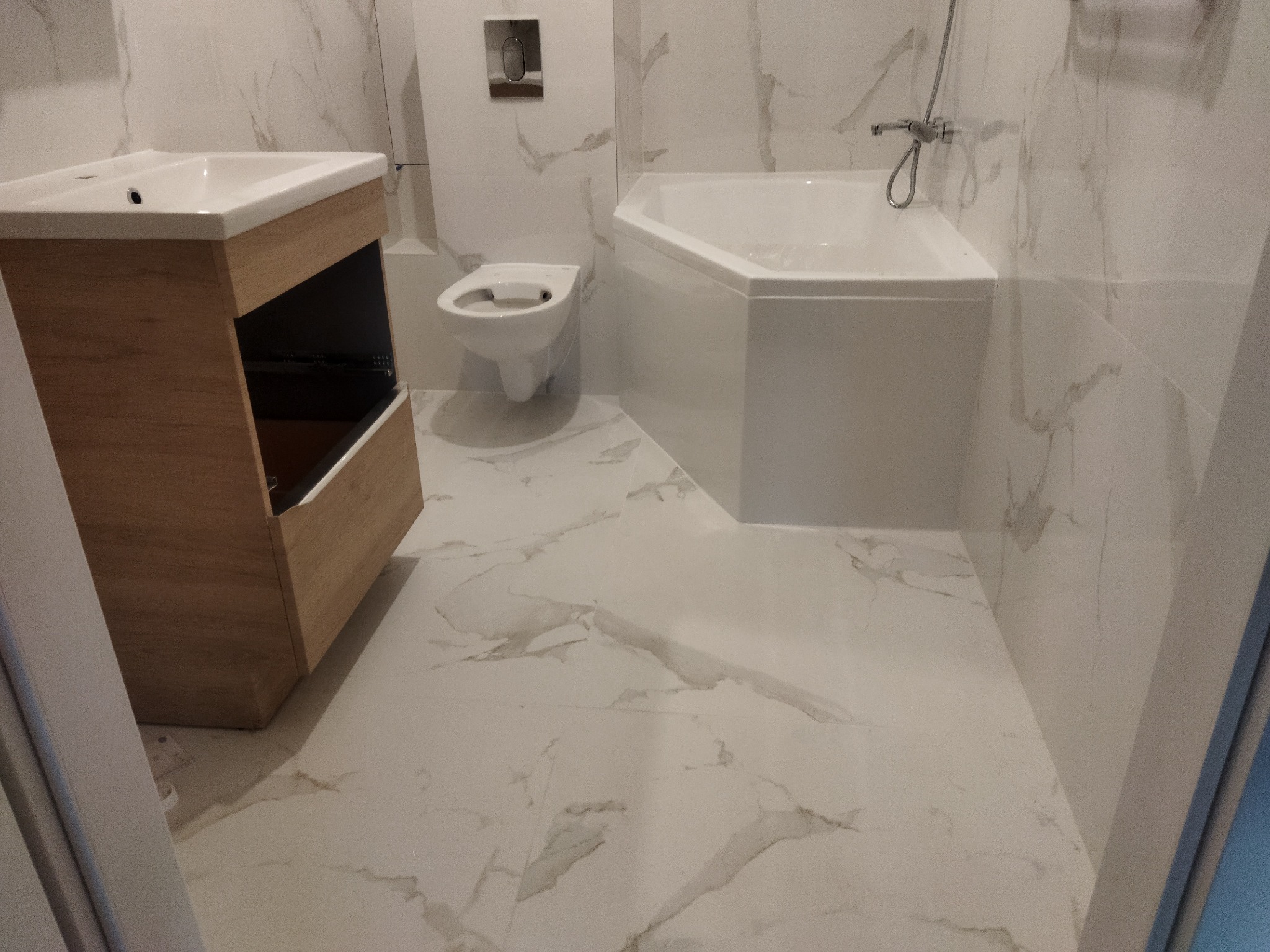 Nowoczesna łazienka z białymi marmurkowymi płytkami, narożną wanną, podwieszaną toaletą i szafką pod umywalką z drewnianym frontem.