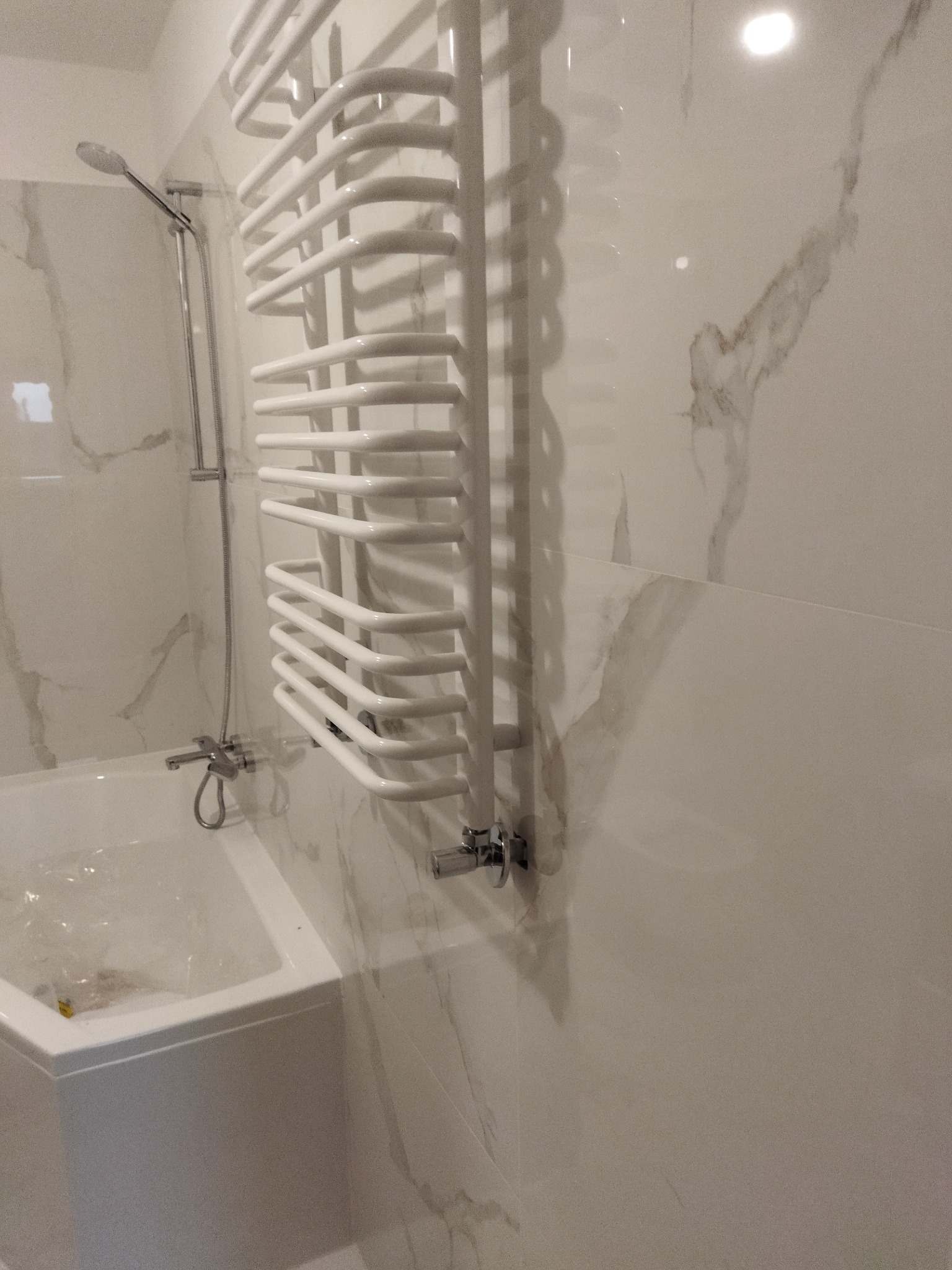 Nowa łazienka z białymi marmurkowymi płytkami, grzejnikiem drabinkowym i wanną z folią ochronną.