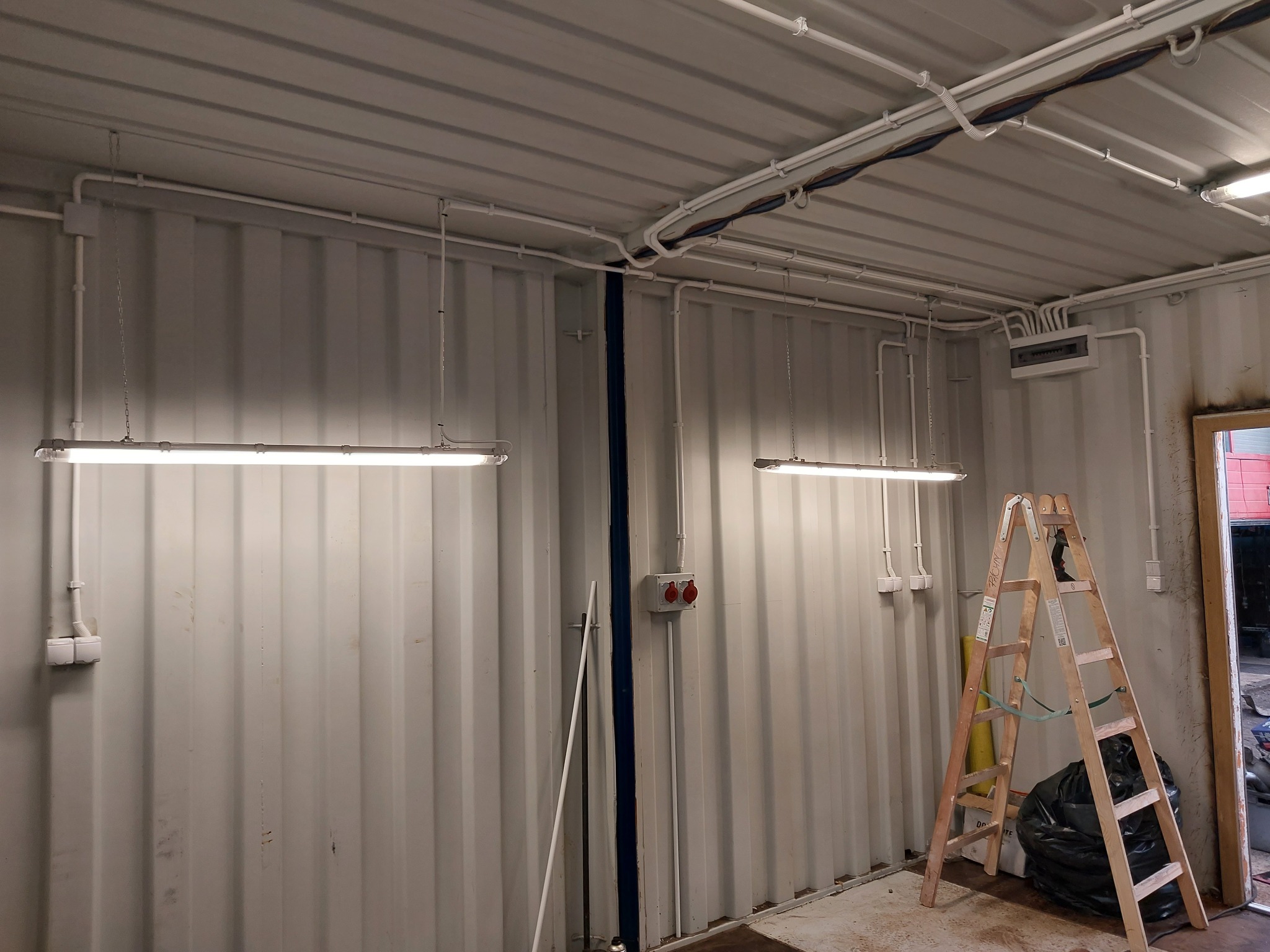 Wnętrze kontenera z widocznymi, nowo zamontowanymi lampami liniowymi LED, instalacją elektryczną natynkową w białych rurkach PCV, skrzynką rozdzielczą i drabiną drewnianą z workiem na śmieci u...