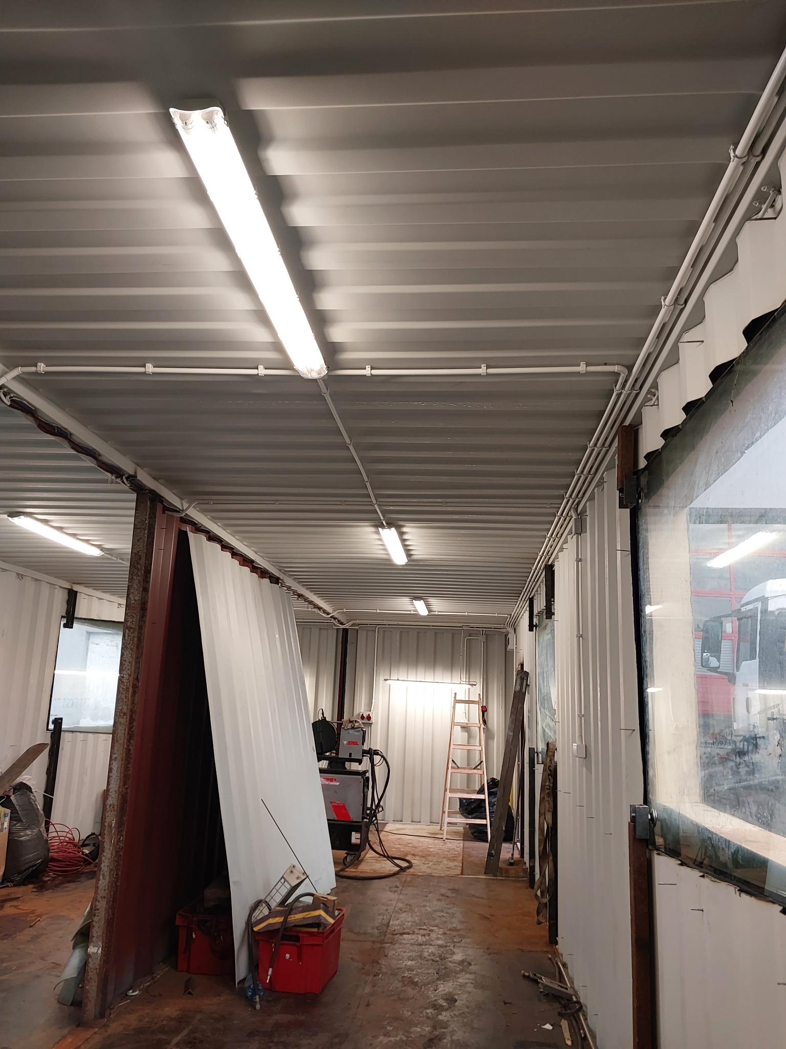 Wnętrze warsztatu w kontenerze z widocznymi przewodami elektrycznymi na suficie i ścianach, oświetleniem jarzeniowym, spawarką, drabiną i innymi narzędziami.
