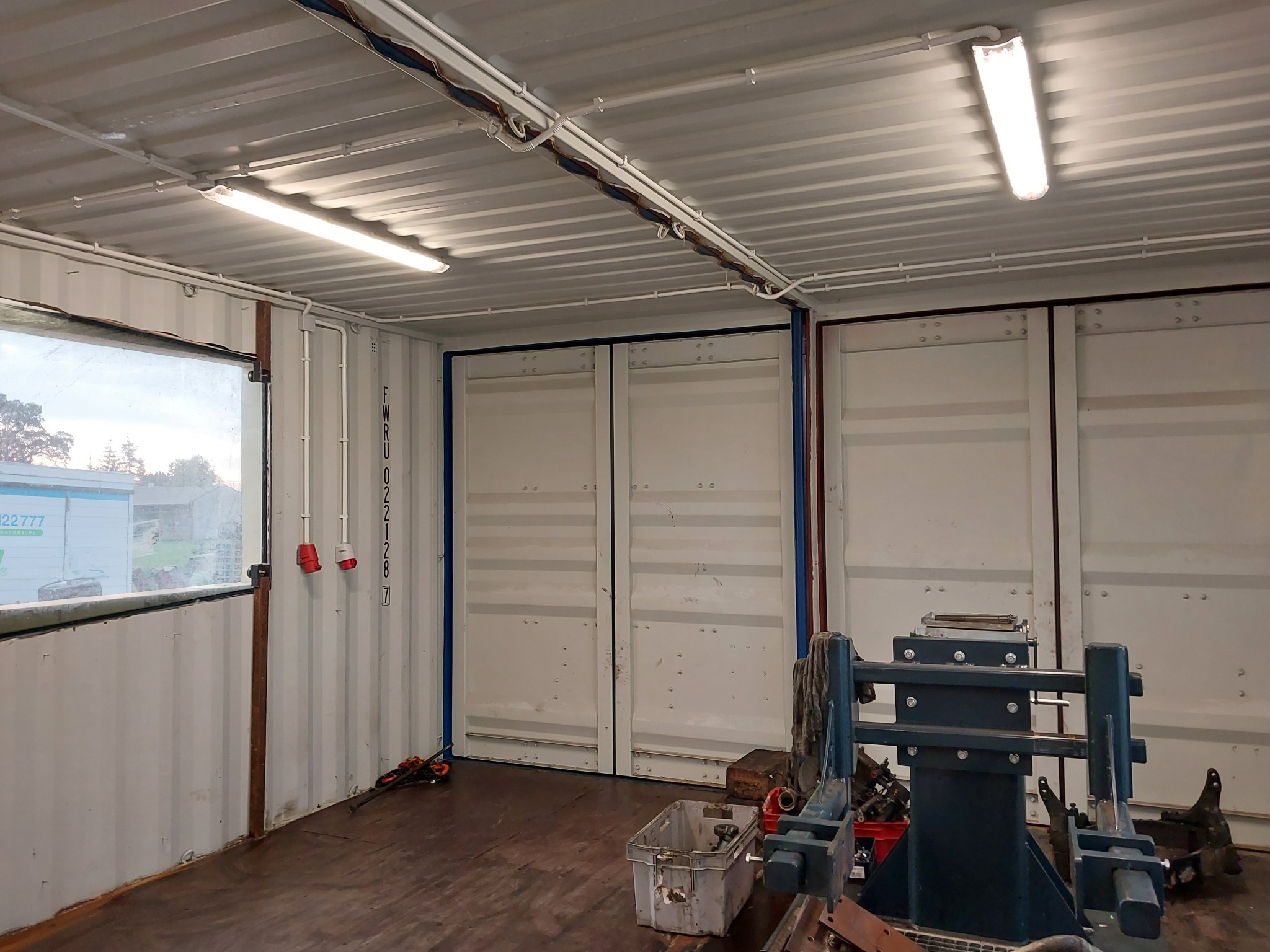 Wnętrze kontenera z widocznymi przewodami elektrycznymi prowadzonymi po suficie, oświetleniem jarzeniowym i gniazdami elektrycznymi na ścianie, sugerujące warsztat lub magazyn.