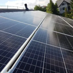 Panele słoneczne zamontowane na ciemnym dachu skośnym, widok z dołu pod kątem, w tle dom jednorodzinny i drzewa.