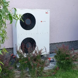 Biała pompa ciepła Saunier Duval zainstalowana przy ścianie budynku w Tomaszowie Lubelskim, widoczna zza krzewów i drzewa płaczącej wierzby, z narzędziami leżącymi obok.