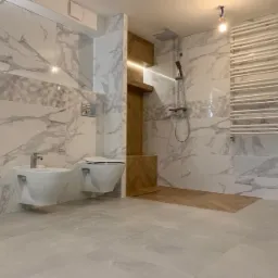 Elegancka łazienka z marmurowymi płytkami na ścianach, wiszącą toaletą i bidetem, drewnianymi akcentami oraz grzejnikiem drabinkowym.