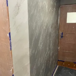 Ściana z efektem dekoracyjnym w odcieniach szarości i bieli, zabezpieczona folią drzwi i fragment ściany podczas prac.