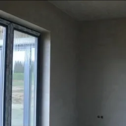 Wnętrze pokoju z oknem, ściany pokryte świeżym, szarym tynkiem, widoczne dwa otwory na gniazdka elektryczne.