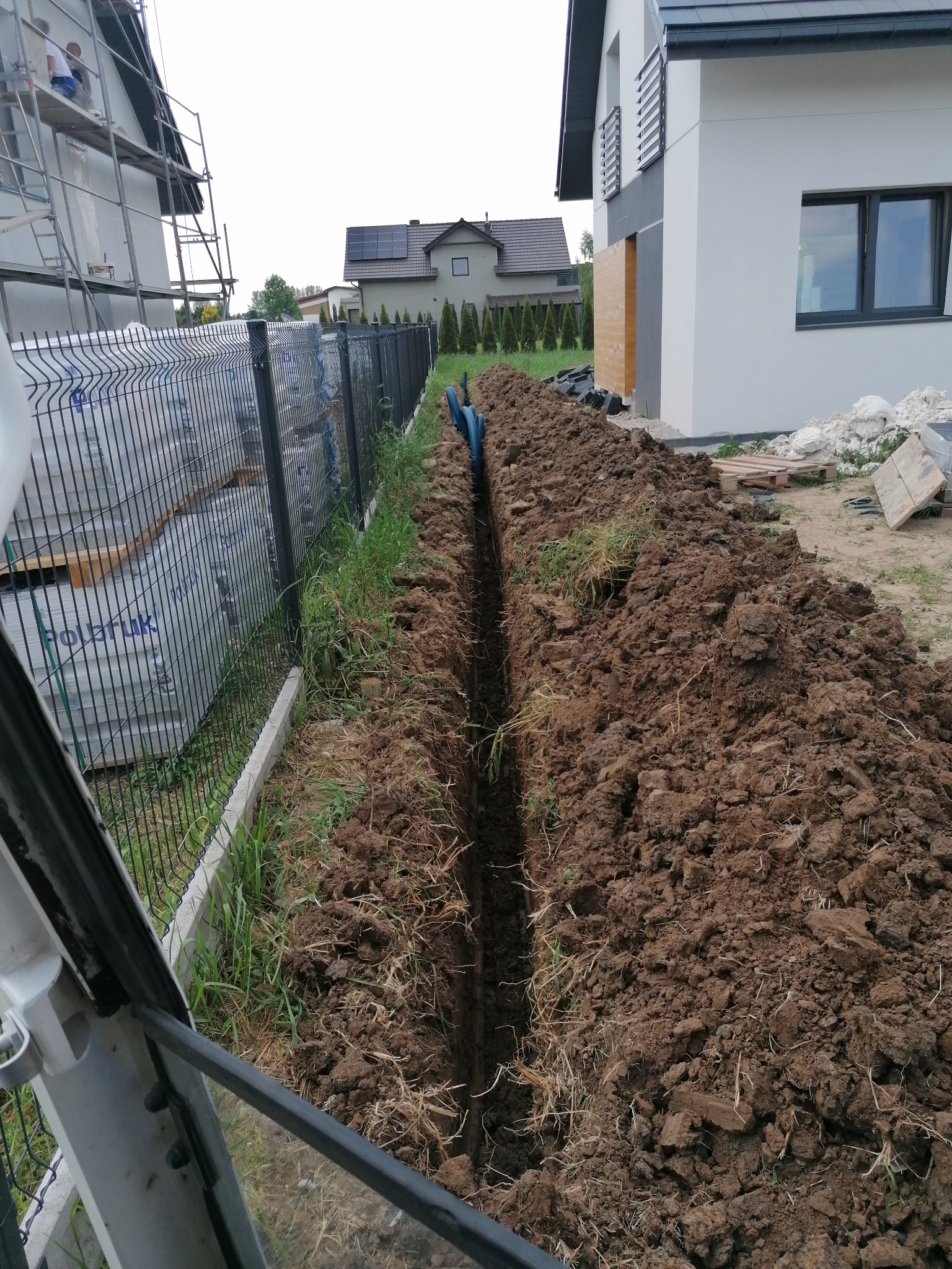 Wąski wykop w ziemi, prawdopodobnie pod instalacje, widoczny z perspektywy operatora maszyny budowlanej, z fragmentem ogrodzenia panelowego po lewej stronie i budynkami w tle.