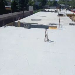 Wylana płyta fundamentowa domu jednorodzinnego z wystającymi prętami zbrojeniowymi, widoczny pracownik w oddali i elementy szalunków.